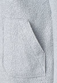 Ljust grått, texturerat tyg med en ficka i triangulär form som lyfter fram en mjuk, lockig textil design.