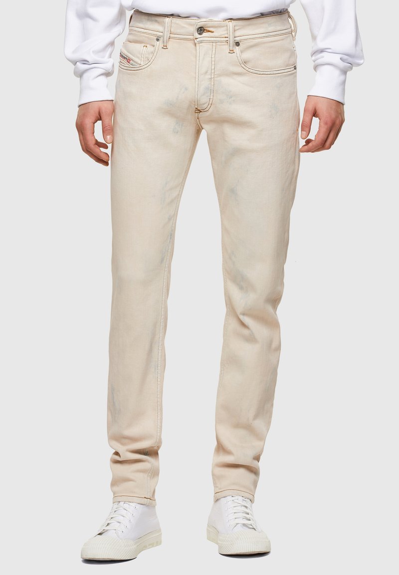 Jeans de mezclilla en un tono beige claro con corte ajustado, que presentan una textura desgastada y un clásico diseño de cinco bolsillos. Combinados con zapatillas blancas.