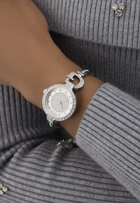 Ezüstóra kör alakú számlappal, amelyet gyöngyházakkal díszítettek és texturált szíjjal rendelkezik. Különleges csattal van ellátva, és minimalista dizájnnal rendelkezik.