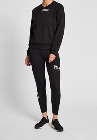 Sweat-shirt noir avec logo blanc sur la poitrine, associé à un legging noir avec texte blanc et accents latéraux. Porté avec des chaussures de sport noires.