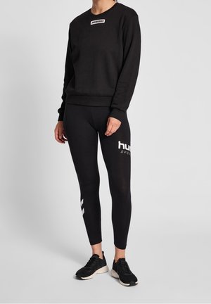 Sweat-shirt noir avec logo blanc sur la poitrine, associé à un legging noir avec texte blanc et accents latéraux. Porté avec des chaussures de sport noires.