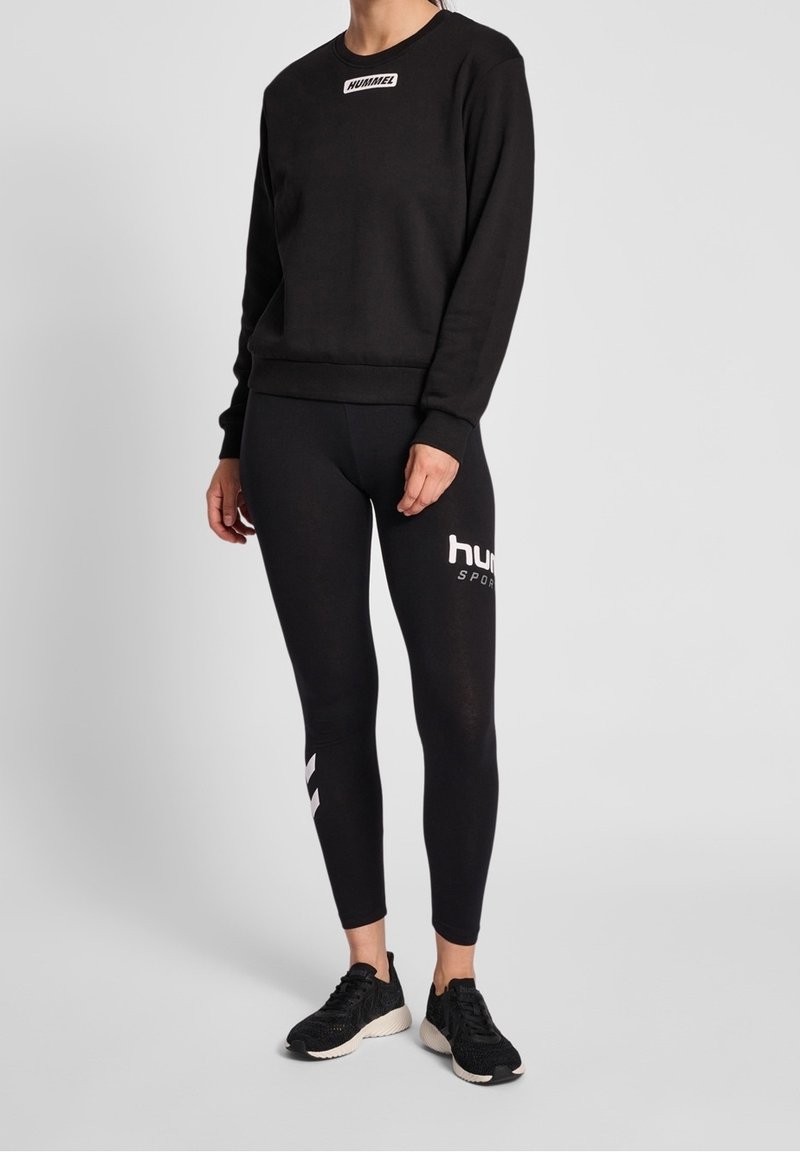 Sweat-shirt noir avec logo blanc sur la poitrine, associé à un legging noir avec texte blanc et accents latéraux. Porté avec des chaussures de sport noires.