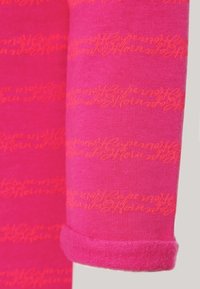 Jasnopinkowa bluzka z długim rękawem z powtarzającym się pomarańczowym wzorem w formie napisu. Miękka tkanina o gładkiej teksturze i wykończeniu mankietów w formie rolki.