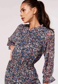 Blumenbluse aus leichtem, durchsichtigem Stoff. Merkmale sind ein Rüschensaum, lange Puffärmel und ein elastischer Taillenbund in Dunkelblau, Pink und Weiß.