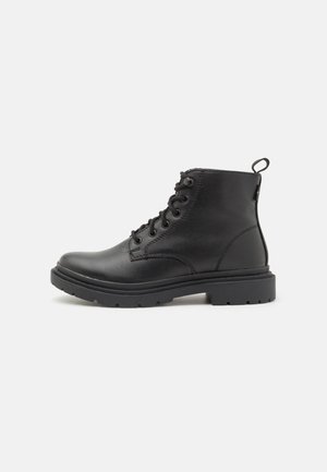 Bottine en cuir noir avec semelle épaisse, à lacets à l'avant et avec une languette à l'arrière, présentée en vue de profil sur fond uni.