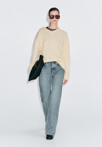 Vrouw die loopt, gekleed in een oversized beige trui, lichtblauwe jeans, zwarte laarzen, grote zonnebril en een zwarte clutch vasthoudt tegen een witte achtergrond.
