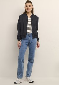 Navy Bomberjacke mit rundem Halsausschnitt, kombiniert mit einem schwarz-weißen gestreiften Hemd, hellblauen Jeans und weißen Sneakers.