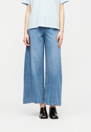 Persona che indossa jeans a gamba larga color azzurro chiaro, stivaletti neri lucidi alla caviglia e una maglietta a maniche corte azzurro chiaro, in piedi davanti a uno sfondo semplice.