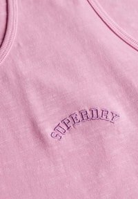 Jasnoróżowy top na ramiączkach z okrągłym dekoltem, z napisem "SUPERDRY" wyszytym w ciemniejszym odcieniu nad środkiem. Gładka tekstura.