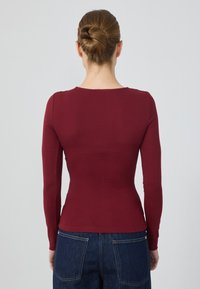 Top bordeaux a maniche lunghe con design slim, scollatura rotonda e texture liscia, abbinato a jeans a gamba larga blu scuro.