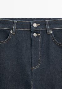 Granatowe dżinsy z denimu z dwoma srebrnymi guzikami w talii, kontrastowymi żółtymi szwami i klasycznym pięciokieszeniowym krojem.