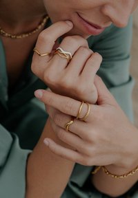 Inele de aur în diverse modele: trei benzi ondulate, un accent alb și un model răsucit de frânghie. Mâinile odihnindu-se pe o ținută verde.