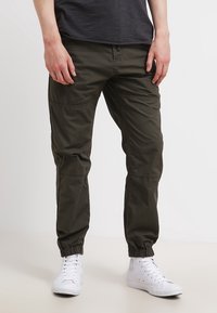 Khaki kalhoty v olivově zelené látce, s elastickými manžetami, bočními kapsami a volným střihem. Nenošeny s bílými teniskami.