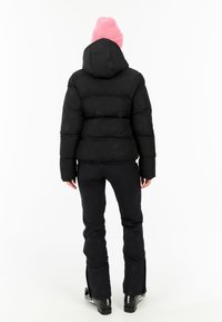 Zwarte pufferjack met sterpatroon, capuchon en elastische zoom, gecombineerd met zwarte broeken met ritsen bij de enkel, gedragen met een roze gebreide muts.