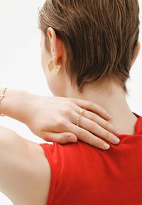 Pendientes en espiral de oro, una pulsera de cruz de oro y un anillo delicado se llevan con una blusa roja. El cabello es corto y texturizado.