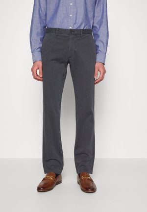 Mężczyzna w szarych prostych spodniach, niebieskiej koszuli na guziki w wzory oraz brązowych skórzanych mokasynach stoi na jednolitym tle.