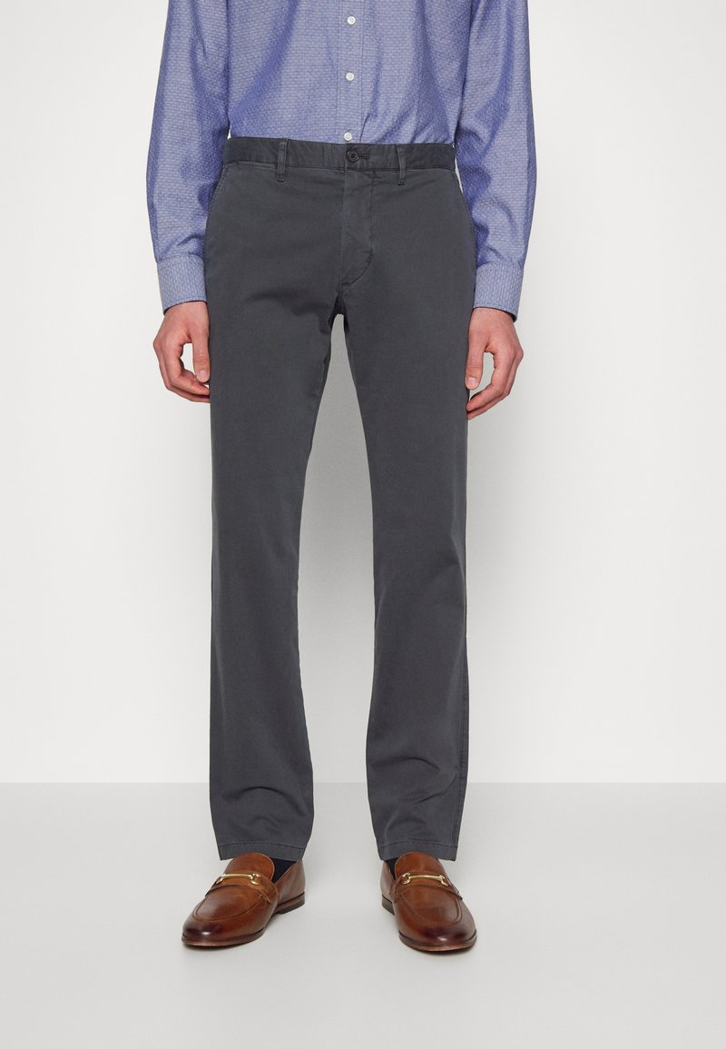 Homme portant un pantalon gris droit, une chemise à motifs bleue boutonnée, et des mocassins en cuir marron, debout devant un fond uni.