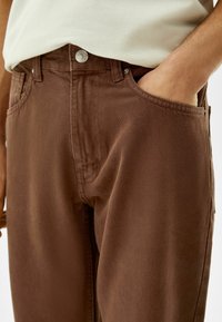 Pantalones de algodón marrón con una textura suave, que cuentan con un bolsillo delantero y un cierre de botón metálico. El diseño es simple y moderno.