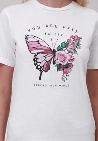 Bílý tričko s velkým růžovým motýlem a květinovým vzorem. Text zní "JSTE SVOBODNÍ létat" nahoře a "ROZPÍREJTE SVÁ KŘÍDLA" dole.