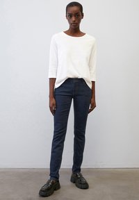 Witte katoenen blouse met lange mouwen gecombineerd met donkerblauwe slim-fit jeans en stevige zwarte schoenen, staande op een effen achtergrond.