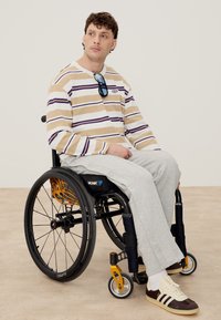 Νεαρός άνδρας καθισμένος σε αναπηρικό καροτσάκι, φοράει μπεζ και μωβ ριγέ πουλόβερ, ανοιχτό γκρι παντελόνι, λευκές κάλτσες και αθλητικά παπούτσια με γυαλιά ηλίου κρεμασμένα στο πουλόβερ.