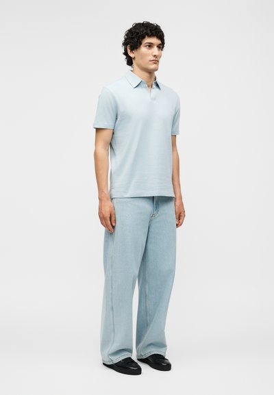 Άνδρας με σγουρά μαλλιά, που στέκεται με το πρόσωπο προς τα εμπρός, φοράει ανοιχτό μπλε πόλο, φαρδιά ανοιχτόχρωμα τζιν και μαύρα παπούτσια σε λευκό φόντο.