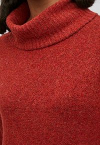 Pull en tricot rouge avec un col roulé côtelé haut. La texture est douce et chaude, avec un motif chiné subtil.