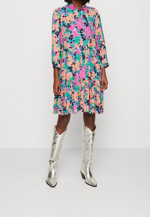 Femme portant une robe colorée à motifs floraux avec manches longues et des bottes de cow-boy argentées montant jusqu'aux genoux, debout sur un fond uni clair.