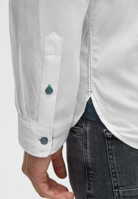 Langärmliges weißes Hemd aus strukturiertem Stoff, mit geknöpften Manschetten, die blaue und grüne Akzente aufweisen, und einem leicht abgerundeten Saum. Graue Jeans sind teilweise sichtbar.