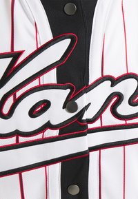 Maglia da baseball con strisce verticali bianche e nere. Presenta lettering ricamato in nero e rosso e bottoni a pressione neri. Tessuto testurizzato.
