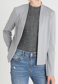 Jasnoszary blazer o luźnym kroju, z otwartym przodem i subtelną teksturą materiału, nałożony na ciemnoszary top prążkowany i zniszczone dżinsy.