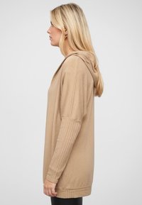 Beige hoodie met geribbelde lange mouwen, zachte textuur, ontspannen pasvorm en een iets gebogen zoom, gedragen over zwarte leggings.