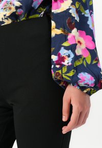 Hånden hviler mod sorte bukser, iført en langærmet bluse med en marineblå baggrund og livligt blomsterprint.
