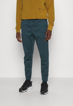 Person trägt ein senfgelbes Langarmshirt und dunkel-türkise Nike-Sweathosen kombiniert mit schwarz-weißen Nike-Sneakers, steht auf weißem Boden.