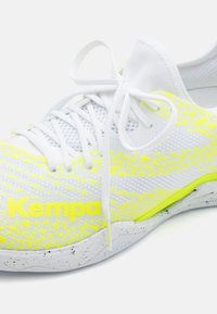 Scarpa sportiva bianca con superficie testurizzata, dettagli in giallo neon e suola grigia chiazzata, dotata di un design tradizionale con lacci.