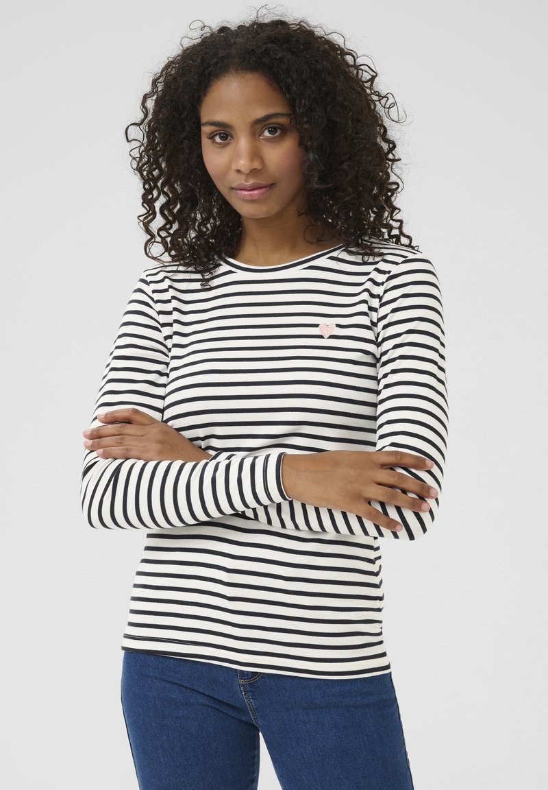 Femme aux cheveux bouclés portant une chemise blanche à manches longues avec des rayures noires et un jean bleu, debout les bras croisés.