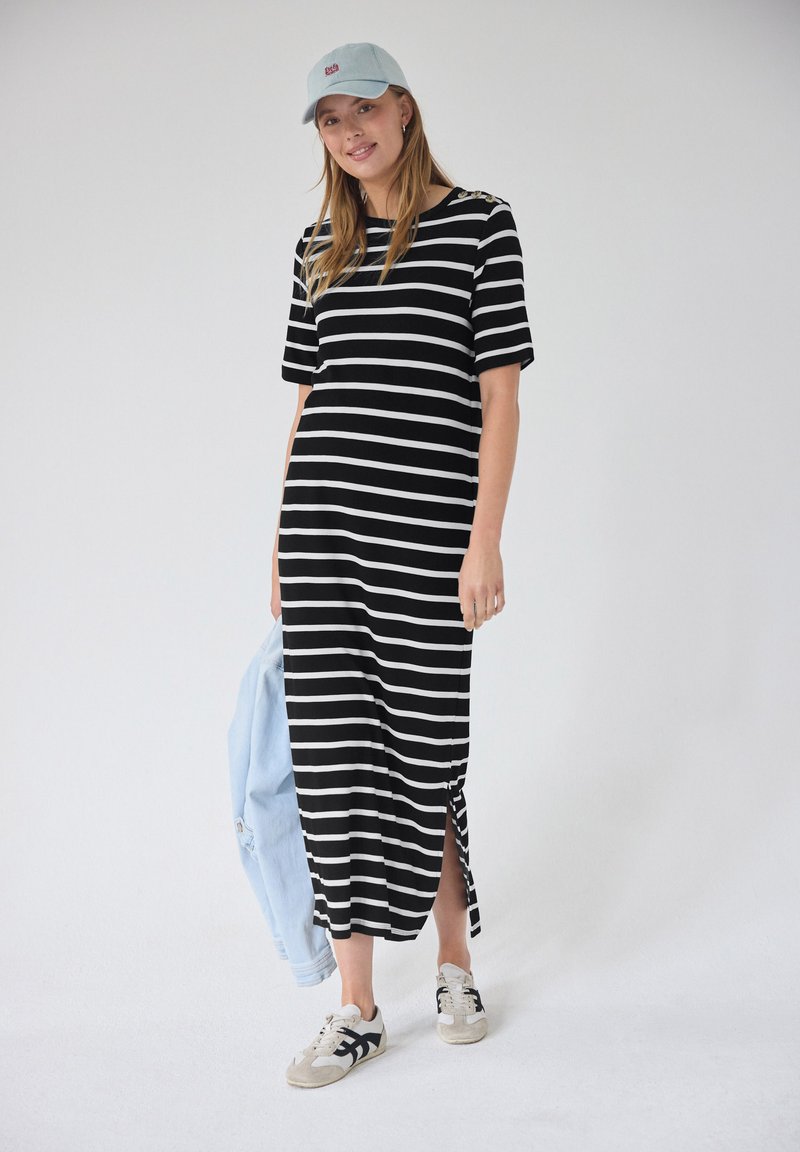 Femme en robe midi rayée noir et blanc, casquette bleu clair, baskets blanches, tenant une veste bleu clair sur fond uni.
