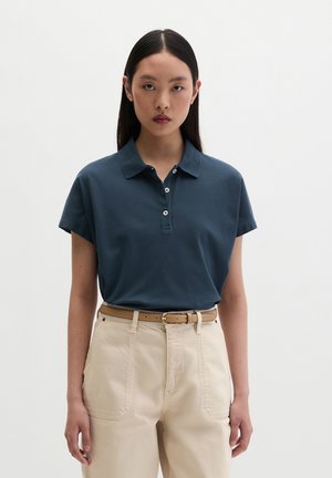 Kobieta o długich czarnych włosach, ubrana w ciemnoniebieską koszulkę polo z krótkim rękawem, wpuszczoną w beżowe spodnie z wysokim stanem i brązowym paskiem.