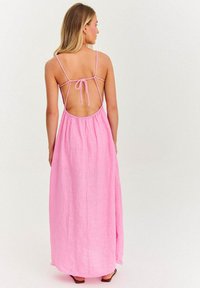 Różowa, lniana sukienka maxi z niskim, otwartym plecami i wiązanymi ramiączkami. Charakteryzuje się swobodnym krojem oraz miękką teksturą. Widoczne płaskie sandały na dole.