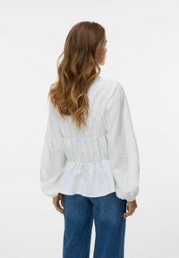 Blusa bianca con texture, maniche lunghe a sbuffo, vita arricciata e fondo fluente, abbinata a jeans in denim blu.