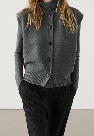 Gray knit cardigan with a high neck, five black buttons, and ribbed hem. Paired with black pinstripe trousers. Minimalistic design.