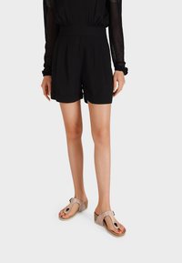 Pantaloni corti neri a vita alta con pinces e orli arrotolati, abbinati a sandali rosa testurizzati con un dettaglio decorativo a loop e suola a zeppa.
