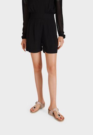 Pantaloni corti neri a vita alta con pinces e orli arrotolati, abbinati a sandali rosa testurizzati con un dettaglio decorativo a loop e suola a zeppa.
