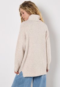 Beżowy sweter dziany z wysokim kołnierzem, ribbowymi mankietami i luźnym krojem. Posiada rozcięty dół oraz miękką, teksturowaną tkaninę.