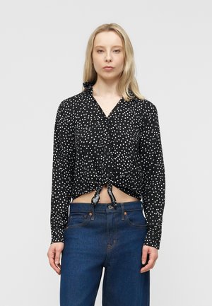 Γυναίκα που φοράει μαύρο πουκάμισο με μακριά μανίκια και άσπρες πουά βούλες, δεμένο στη μέση, και σκούρο μπλε τζιν, μπροστά σε απλό φόντο.