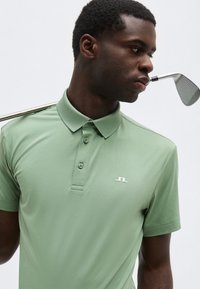 Άνδρας με ανοιχτό πράσινο πόλο πουκάμισο κρατώντας μπαστούνι γκολφ στον ώμο, κοιτάζοντας προς τα πλάγια σε απλό φωτεινό φόντο.