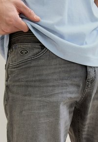 La mano de una persona descansa en la cintura de unos pantalones vaqueros grises, parcialmente cubiertos por una camisa de color azul claro. El entorno es neutral y minimalista.