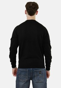 Maglione nero a maniche lunghe con collo giro, polsini a coste, indossato con jeans di denim blu con una toppa e doppia cucitura sulle tasche posteriori.