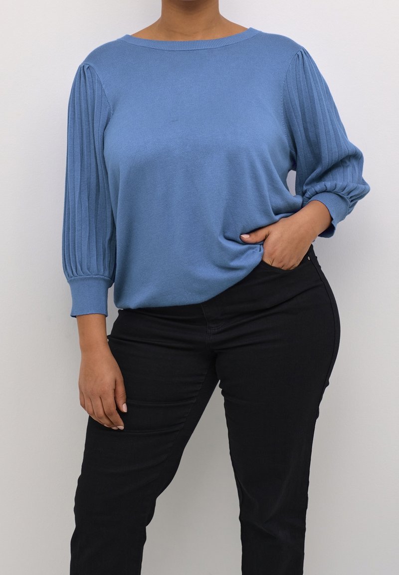 Pull tricoté bleu clair avec manches plissées, encolure arrondie et poignets côtelés, porté avec un pantalon noir ajusté. Texture lisse, coupe décontractée.