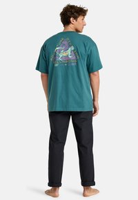 Teal cotton t-shirt with short sleeves featuring a colorful lizard graphic and "Adventure Division" text on the back. Black pants and bare feet.